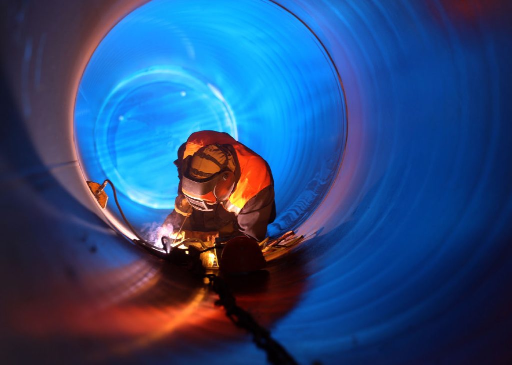 Welder working inside a pipe wearing protective gear with sparks flying.